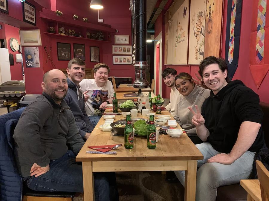 Kubicle team at a hot pot dinner in a red‑walled restaurant, peace signs all round.